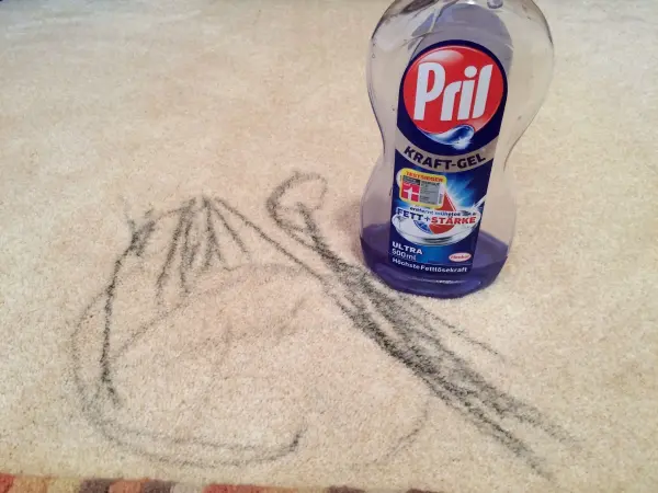 Auf einem hellen Teppich steht eine Flasche Pril, während sich neben ihr bunte Wachsmalstiftfarbe befindet, die entfernt wird.