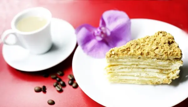 Eine Schichttorte Napoleon liegt auf einem weißen Teller neben einer Tasse Kaffee, umgeben von Kaffeebohnen und einer lila Blume.