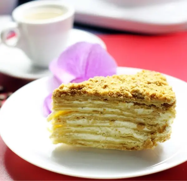 Eine Schichttorte Napoleon liegt auf einem Teller, während eine Tasse Kaffee daneben steht, umgeben von einer eleganten Umgebung.