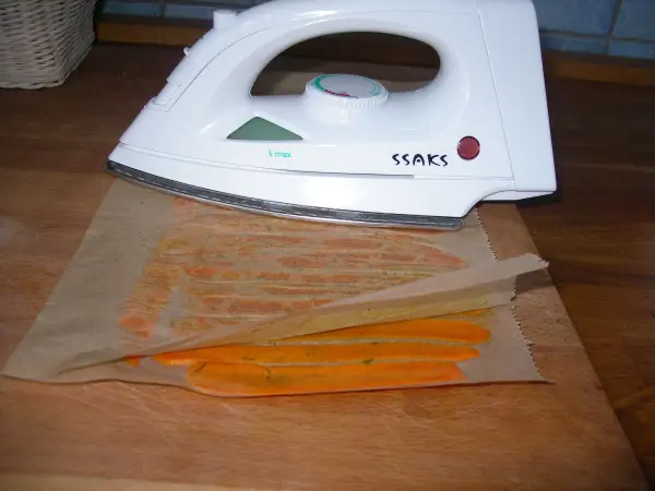 Ein Bügeleisen erhitzt Möhrenchips auf einem Backpapier auf einer Holztischplatte in einer hellen Küche.