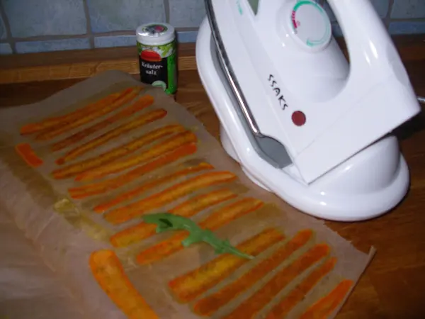Auf einem Holztisch liegen Möhrenchips, die mit einem Bügeleisen bearbeitet werden, während Gewürze daneben stehen.