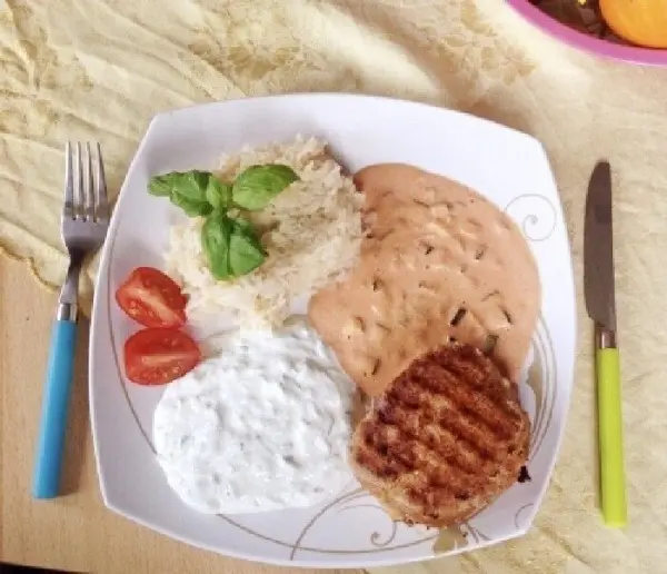 Ein saftiges griechisches Hacksteak liegt auf einem Teller, ergänzt durch Reis, Tomaten und eine Joghurtsoße im Hintergrund.