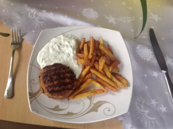 Ein saftiges Hacksteak liegt auf einem weißen Teller, begleitet von knusprigen Pommes und einer weißen Soße.