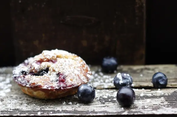 Gesunde Muffins mit Heidelbeeren liegen auf einem rustikalen Tisch, umgeben von frischen Beeren und Puderzucker.