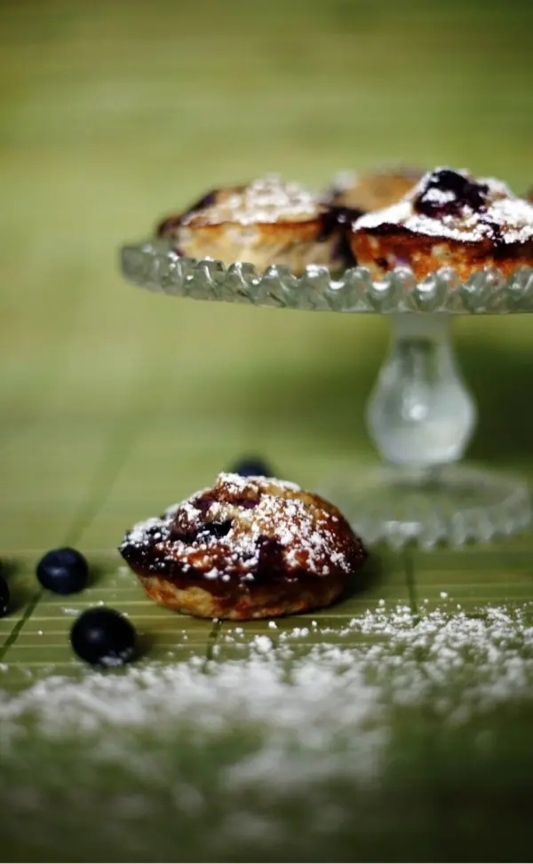 Gesunde Muffins liegen auf einem eleganten Glasstand, umgeben von Heidelbeeren und Puderzucker auf einer grünen Fläche.