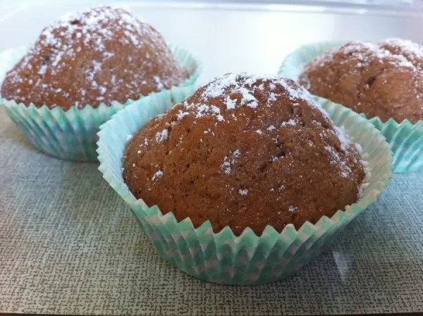 Drei Bananen-Schoko-Muffins liegen in hellblauen Papierförmchen auf einer neutralen Oberfläche und sind mit Puderzucker bestäubt.