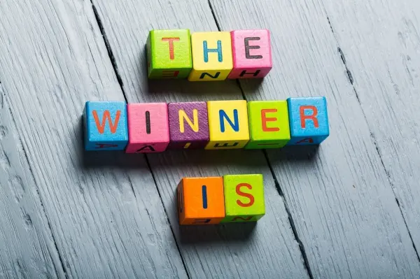 Bunte Buchstaben auf einem Holztisch bilden den Satz "THE WINNER IS", umgeben von einem neutralen Hintergrund.