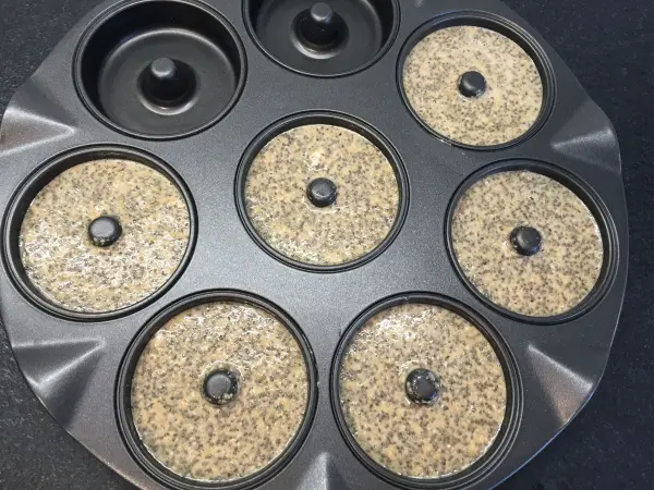 Eine Backform für Chia Donuts mit mehreren runden Vertiefungen, gefüllt mit einer dunklen Teigmischung, steht auf einer dunklen Oberfläche.