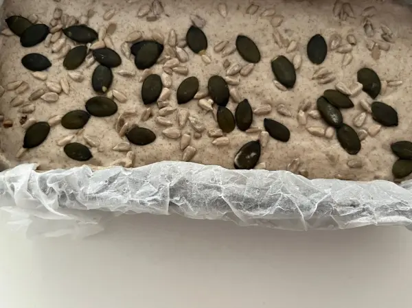 Eine mehlig-braune Teigmischung mit Kürbiskernen und Sonnenblumenkernen in einer Backform ruht für das Brotbacken.
