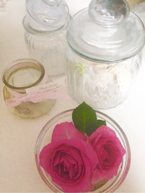 Zwei rosa Rosen liegen in einer Glasschale, umgeben von transparenten Gläsern auf einem hellen Tisch.