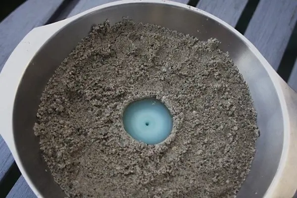 Eine blaue Sandkerze steht in einer Schüssel, umgeben von sandartigem Material auf einer Holzplatte.