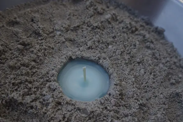 Eine Sandkerze ist in einem Sandhaufen platziert, während der Docht bereit ist, angezündet zu werden. Der Ort wirkt kreativ und handwerklich.