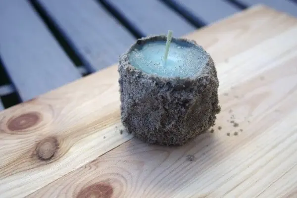 Eine Sandkerze steht auf einem Holzbrett, während sie in der Fertigung sichtbar ist, mit einer unebenen, sandigen Oberfläche.
