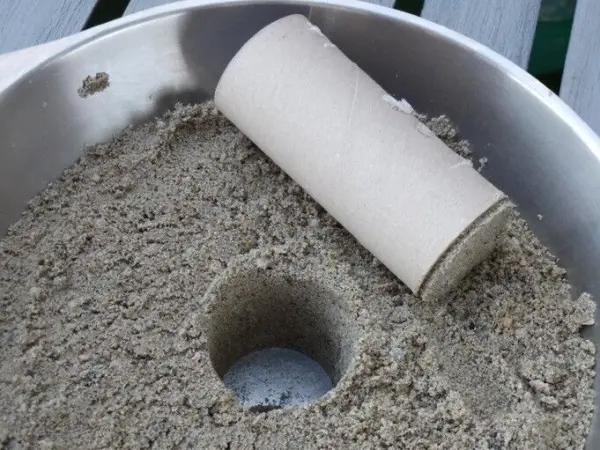 Eine Sandkerze wird vorbereitet, indem ein Pappzylinder in den Sand gedrückt wird. Der Kontext ist eine flache, helle Oberfläche.