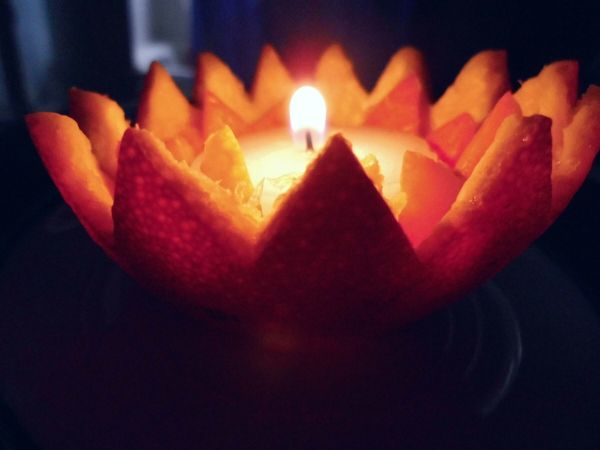 Eine Öllampe in einer Orangenschale brennt sanft, umgeben von auffälligen, leuchtenden Orangenschnitzen in einem dunklen Raum.