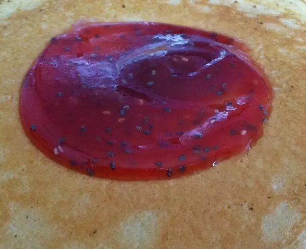Eine rote Erdbeermarmelade mit sichtbaren Chia-Samen ist auf einem hellen, runden Pfannkuchen plaziert.