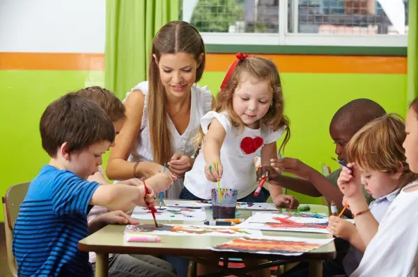 Sechs Kinder und eine Erzieherin malen gemeinsam an einem Tisch in einer kunterbunten Kita-Umgebung.