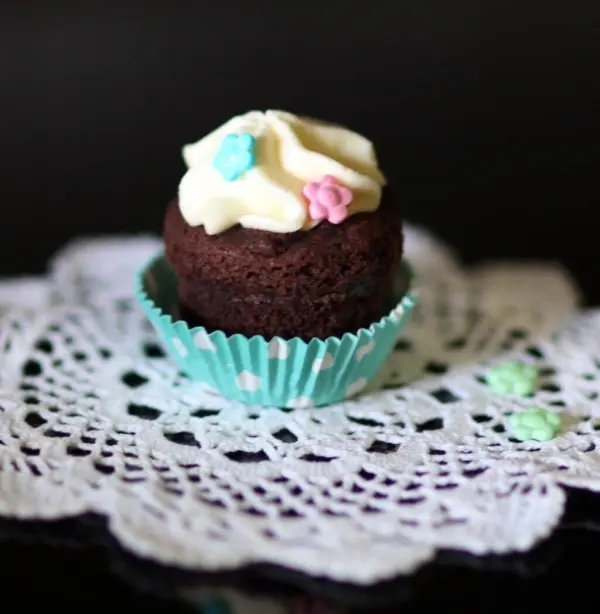 Ein schokoladiger Muffin mit Quarktopping steht auf einem weißen Spitzendeckchen und ist mit bunten Zuckerblümchen dekoriert.