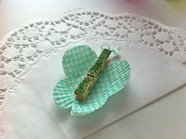 Eine grüne Muffinform ist dekorativ gefaltet und hält eine Holzklammer, die als Schmetterling dient, auf einem weißen Tisch.