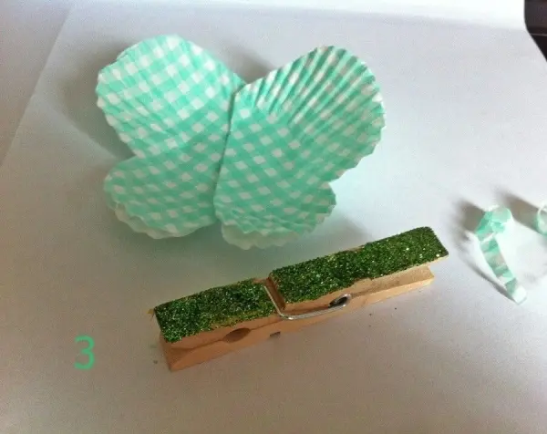 Zwei grüne Muffinformen sind aufgefaltet und bilden die Flügel eines Schmetterlings, daneben eine Klammer mit Glitter.