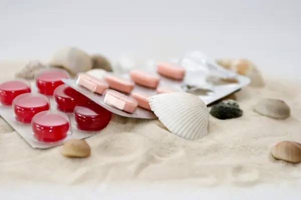 Rote und rosa Tabletten liegen auf Sand, umgeben von Muscheln, was den Kontext eines Urlaubs darstellt und auf eine Reiseapotheke hinweist.