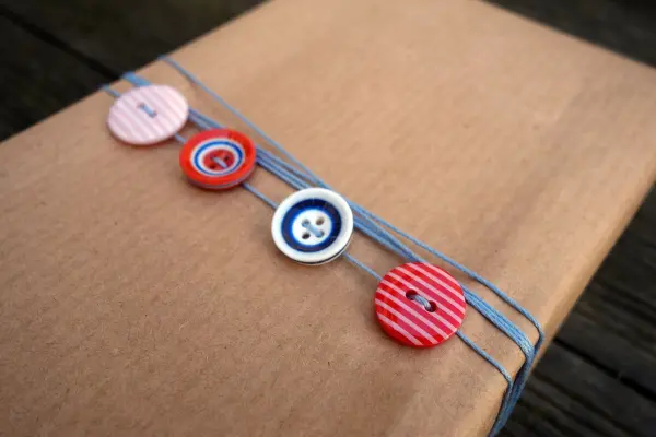 Ein braunes Geschenk ist mit grauem Band und bunten Knöpfen verziert, um eine kreative Verpackung zu gestalten.