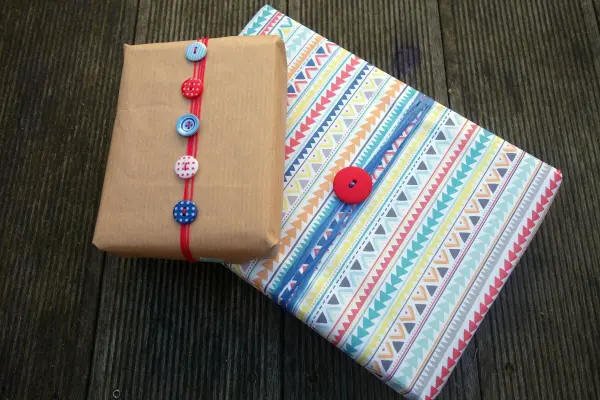 Zwei kreativ verpackte Geschenke mit bunten Knöpfen dekoriert, liegen auf einem rustikalen Holztisch.