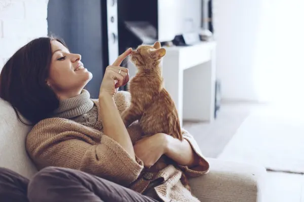 Eine Frau sitzt auf einem Sofa und spielt sanft mit ihrer orangefarbenen Katze in einer gemütlichen Wohnumgebung.