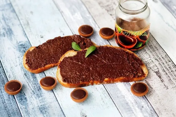 Zwei Brotscheiben mit schokoladigem Toffitella-Aufstrich liegen auf einem rustikalen Tisch, umgeben von Toffifees und einem Glas.