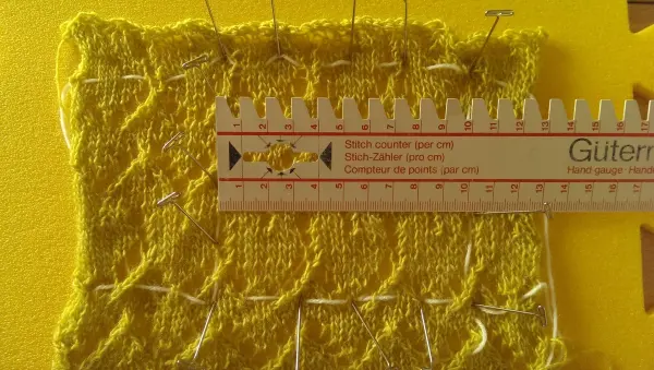 Ein gestricktes Muster in Gelb liegt auf einer gelben Unterlage, während ein Lineal die Maschenprobe misst.