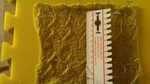 Ein gestricktes, gelbes Maschenprobe-Stück liegt auf einer schaumstoffartigen Unterlage, neben einem Lineal zur Messung.