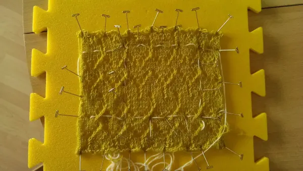 Ein gestricktes Maschenprobe aus gelbem Garn ist auf einer gelben Schaumstoffplatte befestigt, um die Form zu fixieren.