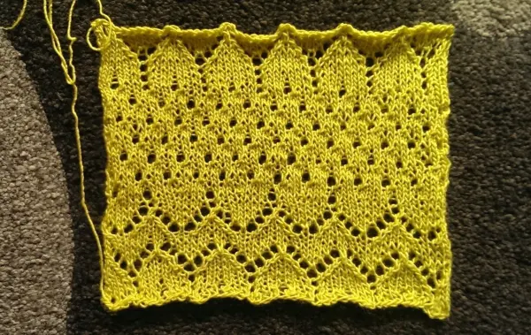 Ein gelbes, gestricktes Tuch zeigt ein regelmäßiges Spitzmuster. Es liegt flach auf einem dunklen Untergrund.