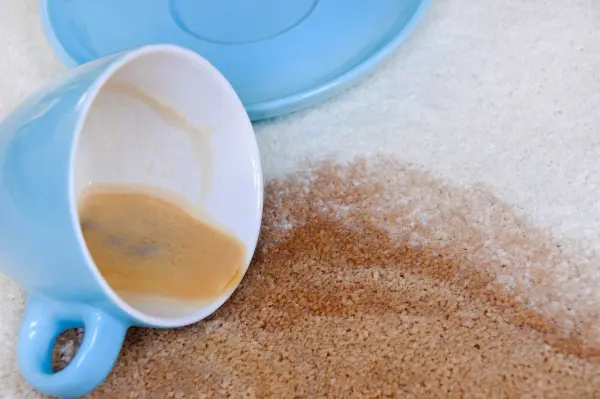 Eine umgefallene, hellblaue Tasse liegt auf einem hellen Teppich, während Kaffeeflecken sich ausbreiten.