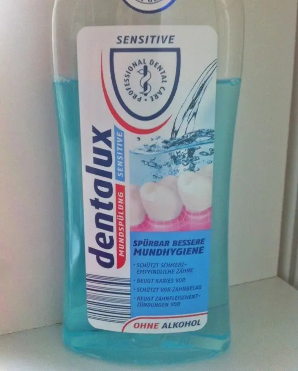 Eine Flasche Zahnspülung mit der Aufschrift „dentalux Sensitiv“ steht auf einem hellen Hintergrund, umgeben von einer Wasserillustration.