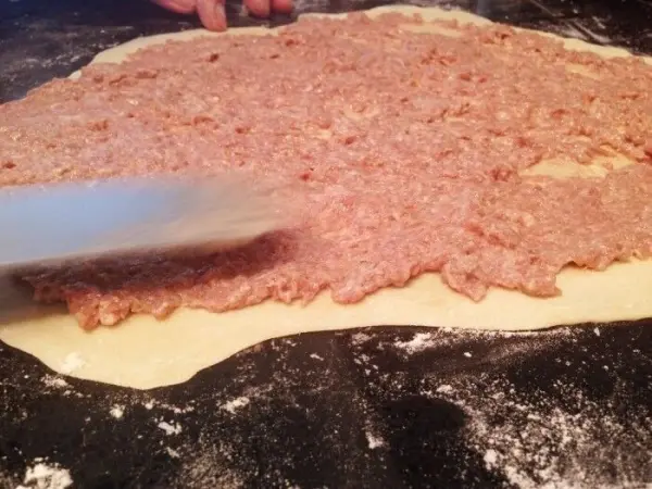 Eine ausgerollte Teigfläche wird mit Hackfleisch gleichmäßig bestrichen, während der Küchenarbeitsplatz gut bearbeitet aussieht.