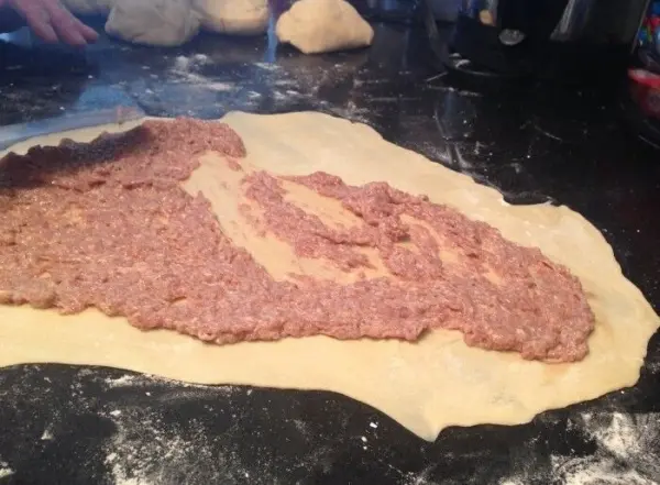 Eine ausgerollte Teigplatte liegt auf einer Arbeitsfläche, mit Hackfleisch gleichmäßig belegt, bereit zum Falten.