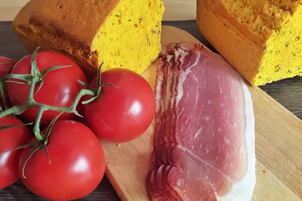 Die goldene Brotscheibe aus rotem Linsenmehl liegt neben frischen Tomaten und dünn geschnittenem Schinken auf einem Holzbrett.