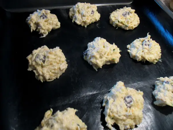 Zucchini-Apfel-Hafer-Cookies liegen ungebacken auf einem schwarzen Backblech, bereit zum Backen in einer Küche.