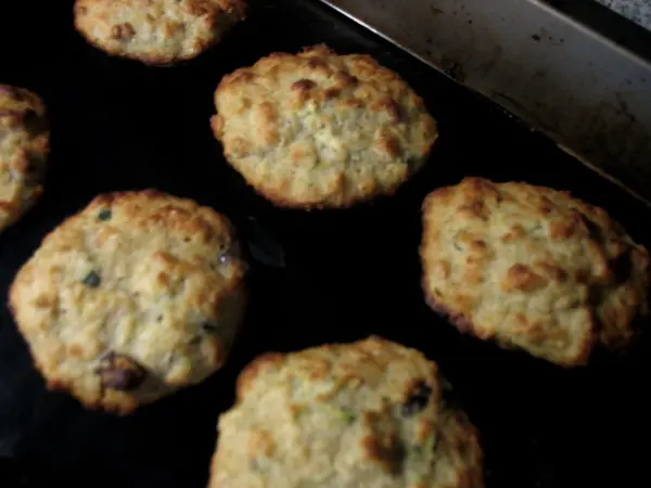Runde Zucchini-Apfel-Hafer-Cookies mit Kokos und Cranberrys liegen gebacken auf einem dunkelgrauen Backblech.