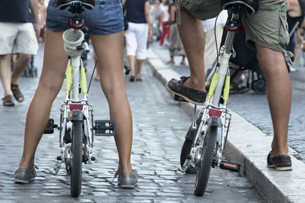 Zwei Personen stehen auf kompakt gefalteten Fahrrädern in einer belebten Straße, während Passanten vorbeigehen.