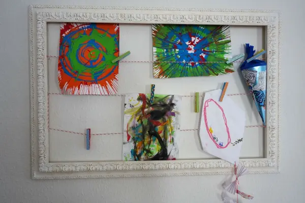 Ein leerer Bilderrahmen dient als kleine Kunstgalerie, in der bunte Kinderzeichnungen auf einer Schnur hängen.