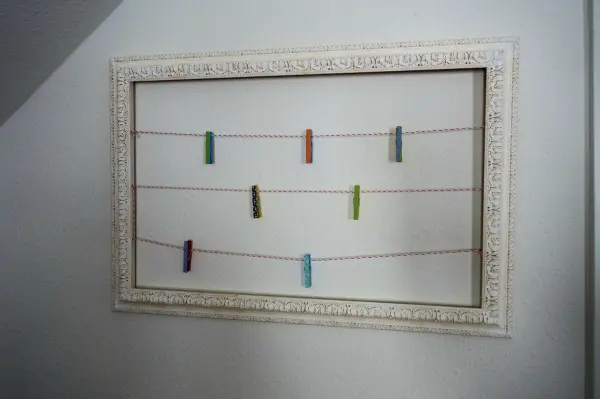 Ein leerer Bilderrahmen hängt an der Wand und zeigt farbige Wäscheklammern, die an Schnüren befestigt sind.
