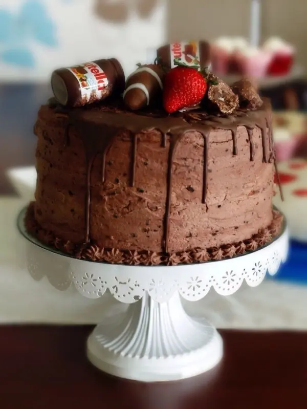 Ein Schokokuchen ist kunstvoll dekoriert mit Nutellagläsern und einer Erdbeere, auf einem eleganten Tortenteller platziert.