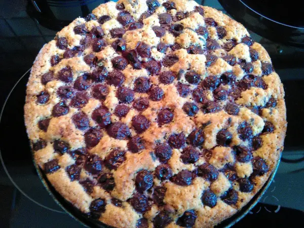 Ein runder Sauerkirschkuchen mit einer goldbraunen, krümeligen Oberfläche steht auf einer dunklen Oberfläche.