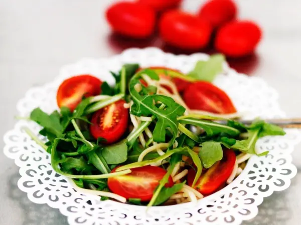 Frischer Salat aus Rucola und Tomaten liegt in einer weißen Schüssel, umgeben von roten Tomaten im Hintergrund.