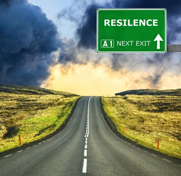 Eine Straße führt in die Ferne, während ein Schild "RESILIENCE (A1 NEXT EXIT)" die Richtung anzeigt. Dunkle Wolken hängen über der Landschaft.