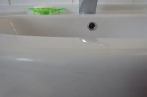 Ein sauberes Waschbecken mit Wasserstrahl, in einem hellen Badezimmer, während ein grünes Reinigungsscheuermittel daneben liegt.