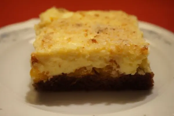 Ein Stück Sägespänekuchen mit Mandarinen auf einem Teller, goldbraun und cremig, vor rotem Hintergrund.
