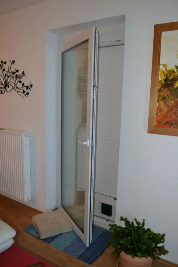 Eine Tür mit einer integrierten Katzenklappe öffnet sich in einen gemütlichen Raum, der mit Pflanzen und Teppichen dekoriert ist.